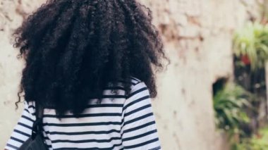 Şehir caddesinde yürüyen güzel bir kadının arka görüntüsü. Sonra kameraya dönüp gülümsüyor. Çekim için poz veren kıvırcık saçlı güzel Afro-Amerikan kadın..