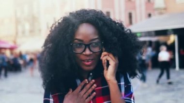 Dışarıda gür kıvırcık saçlı genç değişim öğrencisinin portresi. Ekose gömlekli güzel Afro-Amerikalı kız sokakta dikiliyor ve telefonla konuşuyor..