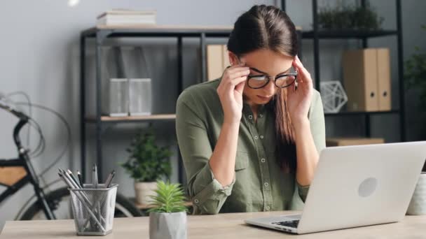Közelkép egy gyönyörű nőről, aki laptopon dolgozik a modern irodában. Fáradt lány leveszi a szemüvegét és masszírozza a halántékát fejfájás miatt.. — Stock videók