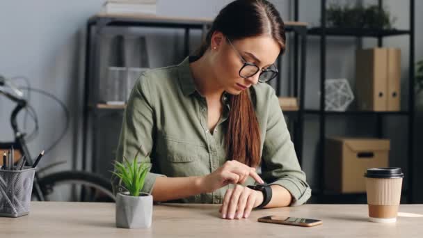 Primer plano de la joven hermosa mujer con gafas usando smartwatch en el trabajo. Mujer atractiva sentada en la oficina configura dispositivo electrónico. — Vídeo de stock