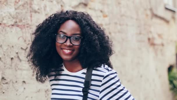 Close up retrato de jovem afro-americana em óculos com cabelo encaracolado. Mulher bonita posando para câmera, sorrindo e alisando o cabelo. — Vídeo de Stock