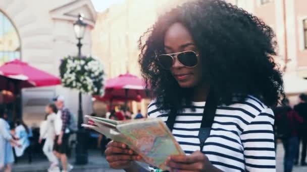 Retrato de una hermosa chica afroamericana mirando el mapa en la calle. Joven turista en gafas de sol con pelo exuberante viajando sola por la ciudad. — Vídeo de stock