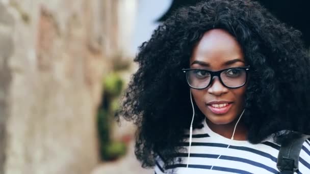 Närbild porträtt av vackra afroamerikanska kvinna stående på gatan och lyssna musik i vita hörlurar. Lycklig flicka med grönskande lockigt hår njuter av musik och leenden vid kameran. — Stockvideo