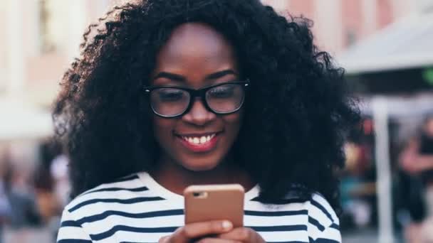 Close up retrato da jovem afro-americana com óculos de pé no meio da rua e segurando o telefone. Menina bonita com cabelo escuro encaracolado olhando para a tela do smartphone fora. — Vídeo de Stock