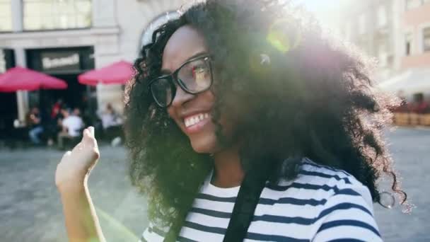 Close up de bela mulher afro-americana de pé no centro da cidade acenando a mão e olhando ao redor. Menina feliz gravação de vídeo na câmera na rua da luz solar. Vista da câmara. — Vídeo de Stock