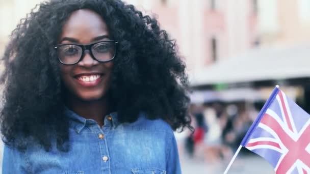 Detailní portrét mladé šťastné výměny studenta se svěží kudrnaté vlasy. Krásná dívka v džínové košili mává britskou vlajkou na ulici. — Stock video