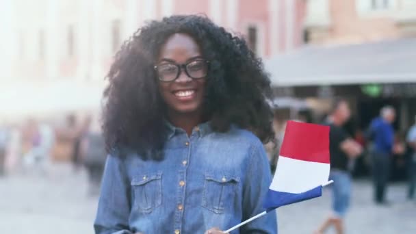 Portrait of young happy exchange student with lush curly hair. Beautiful girl in denim shirt is smiling happily and waving the flag of France on outside. — Stock Video