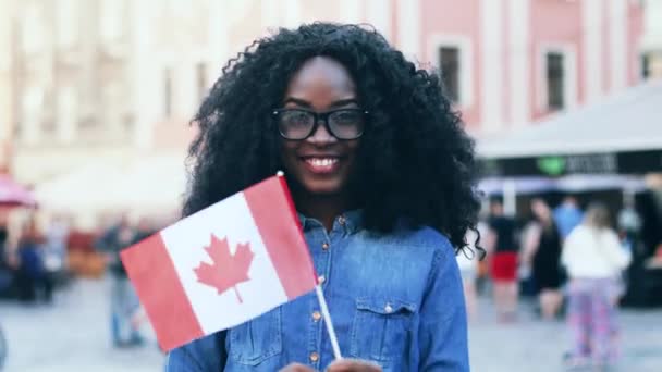 Portret van een jonge vrolijke uitwisselingsstudent met weelderig krullend haar. Mooi meisje in denim shirt lacht vrolijk en zwaait met de vlag van Canada naar buiten. Welkom in Canada.. — Stockvideo