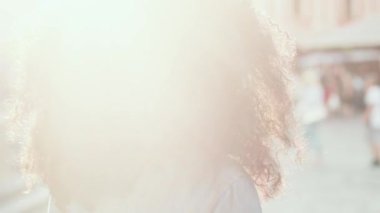 Şehir caddesinde yürüyen güzel bir kadının arka görüntüsü ve sonra kameraya dönüp gün ışığında gülümseyen bir kadın. Kamera, kot gömlekli kıvırcık saçlı güzel Afrikalı Amerikalı kızı takip ediyor..