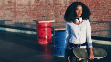 Dışarıda elinde patenle duran kıvırcık saçlı güzel Afro-Amerikan kadın portresi. Beyaz kulaklıklı genç hippi kız kameraya gülümsüyor..