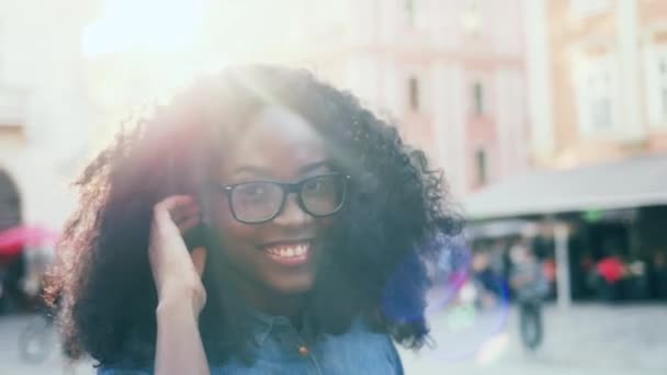 Tutup potret muda african american wanita dalam gelas dengan rambut keriting gelap. Gadis cantik dengan genit tersenyum sambil melihat kamera di bawah sinar matahari di luar. — Stok Video