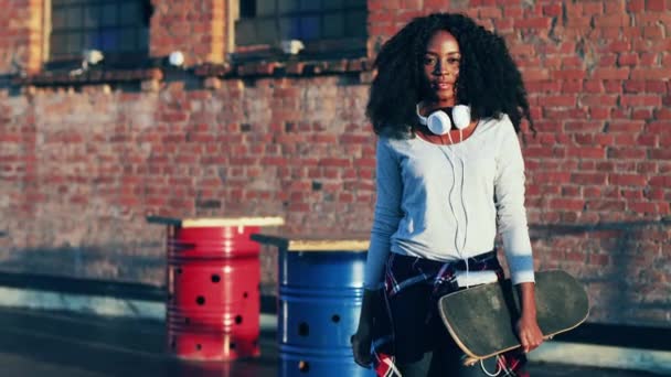 Portret pięknej Afroamerykanki trzymającej deskorolkę w rękach stojącej na zewnątrz. Młoda hipsterka w białych słuchawkach pozuje przed kamerą i uśmiecha się na tle cegły. — Wideo stockowe