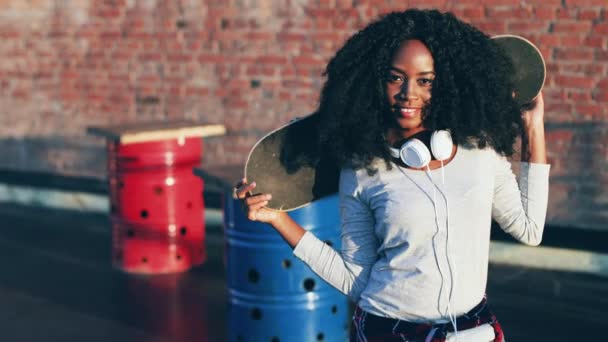 Gros plan de jeune belle femme aux cheveux bouclés avec écouteurs blancs. Hipster fille tenant patiner sur ses épaules et posant pour la caméra à l'extérieur. — Video