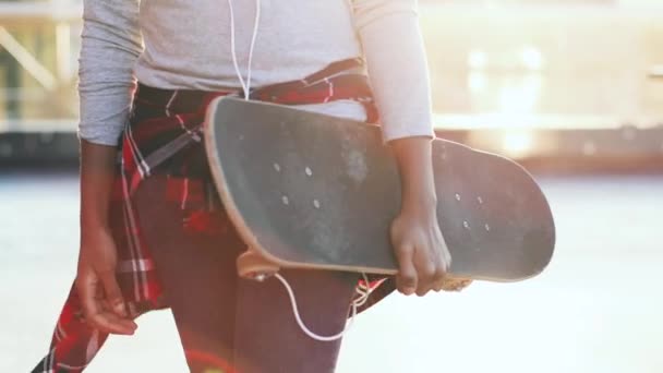 Detailní záběr dívky v džínách s kostkovanou košilí nese skateboard v ruce venku ve slunečním světle. — Stock video