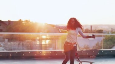 Genç, kıvırcık saçlı Afro-Amerikalı kadın gün batımında çatıda dans ediyor. Güzel kız el kaldırıyor, beyaz kulaklıklarla müzik dinliyor..