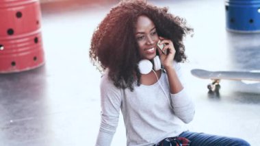 Terasta oturan ve cep telefonuyla duygusal olarak konuşan kıvırcık saçlı neşeli genç bir kadın. Güzel Afrikalı Amerikalı kadın çatıda oturuyor ve mobil cihaz kullanarak iletişim kuruyor..