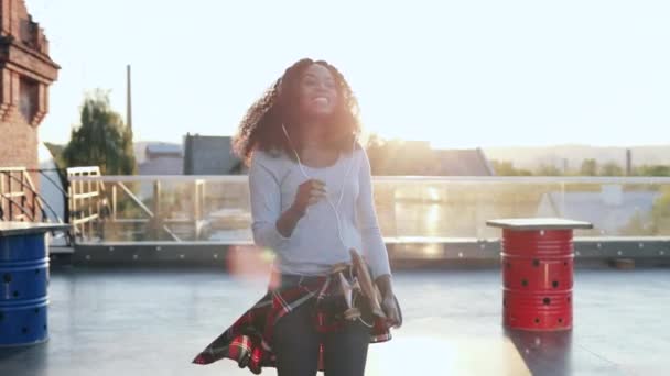 Belle jeune femme afro-américaine aux cheveux bouclés se promène en tenant le patin. Hipster fille marche sur coucher de soleil écouter de la musique dans les écouteurs blancs. — Video