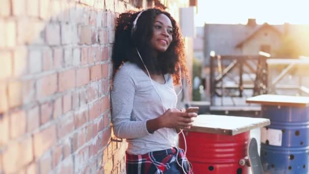 Vacker ung afroamerikansk kvinna med lockigt hår står nära tegelvägg i vita hörlurar. Glada flicka njuter av sin favorit låt på taket av huset i solljuset. — Stockvideo