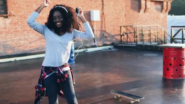 Όμορφη νεαρή Αφρο-Αμερικανίδα με σγουρά μαλλιά που χορεύει στην οροφή του σπιτιού. Ευτυχισμένο κορίτσι με ακουστικά με το τηλέφωνο στο χέρι κινείται προς το ρυθμό της μουσικής στο εξωτερικό. — Αρχείο Βίντεο