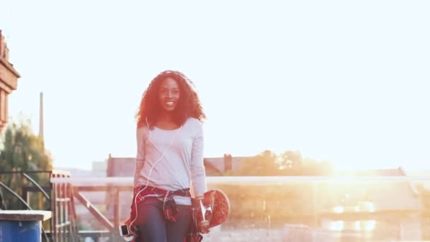 Красивая молодая африканская американка с вьющимися волосами прыгает на фоне кирпичной стены. Веселая девушка слушает музыку в наушниках, держа скейтборд во время прогулки по крыше на закате. — стоковое видео