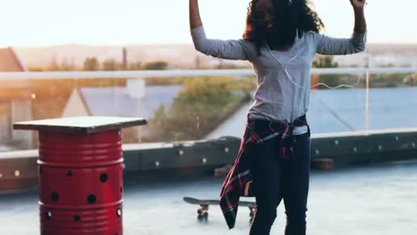 Giovane donna afroamericana felice con i capelli ricci che ballano al tramonto sul tetto. Bella ragazza si muove per la musica in cuffie sulla terrazza. — Video Stock