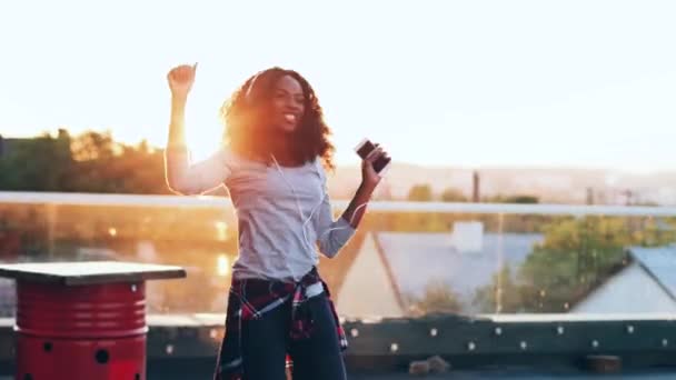 Όμορφη νεαρή γυναίκα ακούει μουσική και χορεύει με ακουστικά στη βεράντα. African American hipster κορίτσι απολαμβάνει ευτυχισμένα ξοδεύουν χρόνο στην κορυφή της οροφής. — Αρχείο Βίντεο