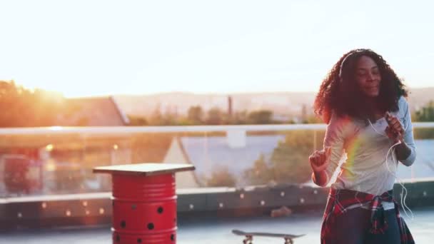 Bella giovane donna con i capelli ricci lussureggianti ascoltare musica e ballare in cuffia sulla terrazza. Afro-americano hipster ragazza felicemente trascorrere del tempo in cima al tetto. — Video Stock