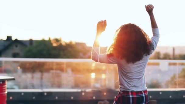 Bella giovane donna con i capelli ricci lussureggianti ascoltare musica e ballare in cuffia sulla terrazza. Ragazza hipster afroamericana in movimento al ritmo della melodia in cima al tetto. Vista posteriore. — Video Stock