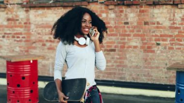 Terasta yürüyen güzel bir kız cep telefonuyla konuşuyor. Genç Afro-Amerikalı hippi kız gür saçlı kaykay taşıyor ve elinde cep telefonu var..