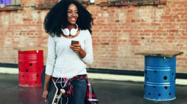 Güzel bir kız büyük bir terasta elinde kaykayla yürüyor. Neşeli Afrikalı Amerikalı kız elinde cep telefonuyla etrafa bakıyor..