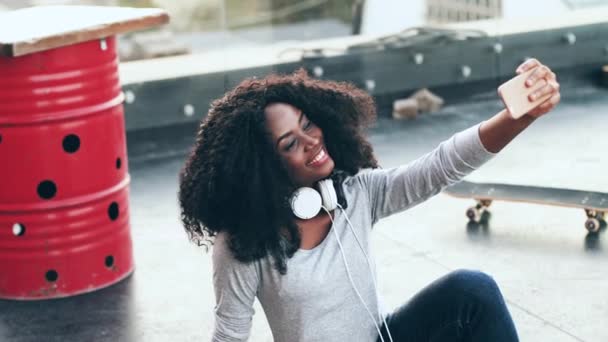 Terasta oturmuş akıllı telefonuyla selfie çeken kıvırcık saçlı neşeli genç bir kadın. Güzel Afro-Amerikalı kadın çatıda oturuyor. Mobil kameraya poz veriyor.. — Stok video