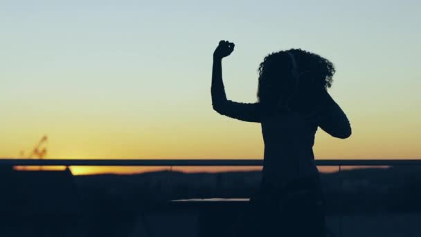 Silueta de niña bailando en el techo al atardecer. Hermosa chica moviéndose a la música en los auriculares fuera. — Vídeos de Stock