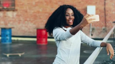 Kıvırcık saçlı neşeli genç kadın terasta duruyor ve akıllı telefonuyla video kaydediyor. Güzel Afro-Amerikalı kadın çatıda dikiliyor. Telefonla konuşuyor.
