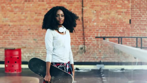 Portrét krásné Afroameričanky s kudrnatými vlasy stojící venku se skateboardem v rukou. Hipster mladá dívka se sluchátky stojící bokem pózovat na kameru s úsměvem. — Stock video