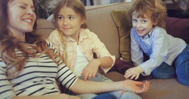 Ευτυχισμένη νεαρή μητέρα που παίζει με τα παιδιά της καθισμένη στον καναπέ. Η μαμά με το γιο και την κόρη περνούν ελεύθερο χρόνο στο σπίτι. — Αρχείο Βίντεο