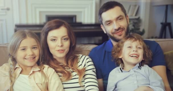 Glückliches Familienporträt von jungen Eltern, Mutter und Vater, die sich mit niedlichen Kindern umarmen. Sohn und Tochter lächeln lächelnd in die Kamera und posieren gemeinsam auf der heimischen Couch. — Stockvideo