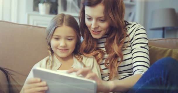 Glückliche junge Mutter und kleine Tochter lächelnd Spaß mit dem digitalen Tablet auf dem Sofa sitzend. Mutter mit Kind hält Tablet in der Hand und blickt auf Bildschirm. — Stockvideo