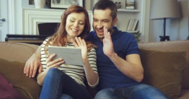 Mutlu evli çift evdeki kanepede otururken tablet kullanarak online arama yapıyor. Videoda konuşan bir kadın saçını düzeltiyor. Karı koca kameraya el sallıyor. Uzak iletişim