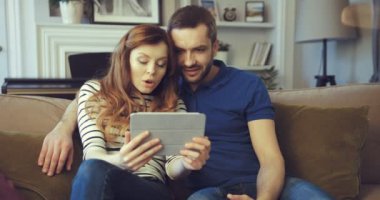 Mutlu evli çift kanepede otururken internette video izliyor. Karısı tableti elinde tutuyor. Güzel bir kadın şaşkınlığını ifade eder..