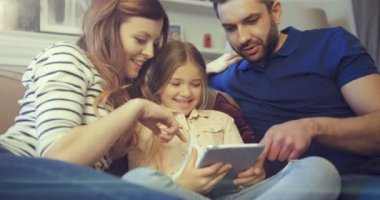 Genç ebeveynler kızlarıyla kanepenin üzerinde elektronik aletle oyun oynuyorlar. Evden tablet okuyup oynayan tatlı bir anaokulu kızı..