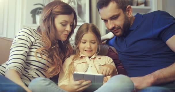 Γονείς με την κόρη παίζουν online παιχνίδι στο tablet στο σπίτι. Ευτυχισμένη μαμά μπαμπάς και η κόρη τους έχουν καλές στιγμές μαζί κάθεται στον καναπέ με gadget. — Αρχείο Βίντεο