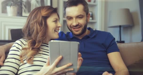 Gelukkig duizendjarig stel genieten van het gebruik van grappige apps op tablet zitten op de bank. Lachende man en vrouw ontspannen thuis tijdens het winkelen online. — Stockvideo