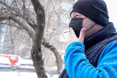Sigara içiyorum. COVID-19 salgını sırasında maskeli bir adam sokakta sigara içiyor. Siyah Coronavirus maskeli bir adam sigara içiyor maskenin içindeki bir delikten sigara içiyor sigara içmenin tehlikeleri