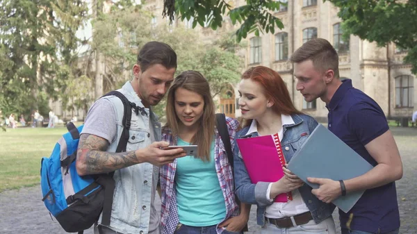 Czterech studentów patrzeć na ekran smartfona na kampusie — Zdjęcie stockowe