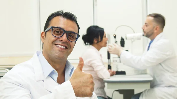 Врач надевает очки и показывает большие пальцы, а на заднем плане другой врач осматривает пациента — стоковое фото