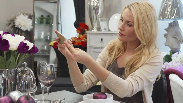 The woman puts the glass in place. Then she corrects the position of the flowers. She takes out the phone and takes a photo of the composition. The woman is sitting at the table