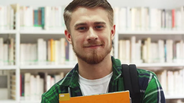 Mladý student s učebnice ve svých rukou stojí v knihovně — Stock fotografie