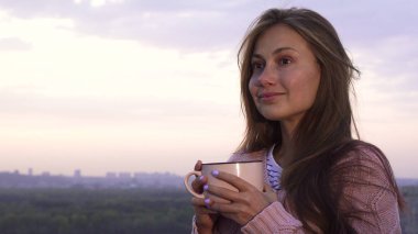 Bir genç kız sıcak bir içki içiyor ve gün batımı sahiptir