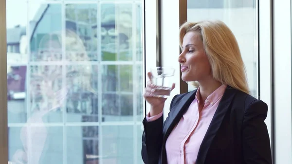 Beautiful woman looks through the window of her office and drinks water — Stock Photo, Image