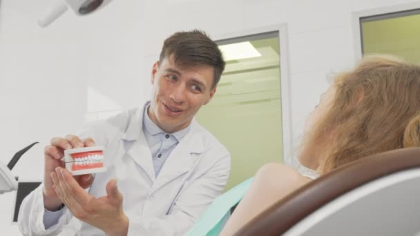 Ragazzina sorridente alla macchina fotografica dentista che tiene i denti modello sul retro — Video Stock